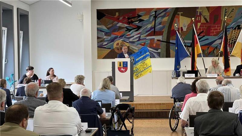 Landrat Kai Seefried stellt das Projekt in einer Kreistagssitzung vor. Er zeigt eine von ukrainischen Frontsoldaten unterschriebene Flagge, die ihm als Dankeschön für die Hilfstransporte überreicht worden war.
