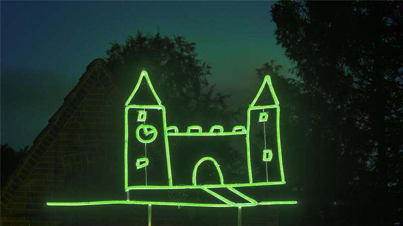 Die grün leuchtende Neon-Lichtskulptur "Luftschloss - Windschief" leuchtet vor der Silhouette von Dach und Bäumen in der Dämmerung