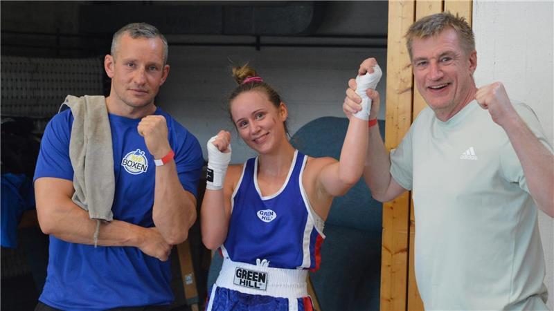 Lilith Nibbe und ihre beiden Trainer posieren mit ihrer Boxerin und strecken die Fäuste nach oben.