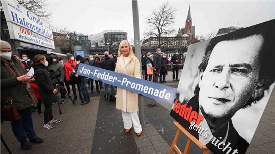 Marion Fedder (M), Witwe von Jan Fedder, hält ein Schild mit dem Text „Jan-Fedder-Promenade“ bei der Umbenennung der Flaniermeile an den Landungsbrücken im Januar 2022.