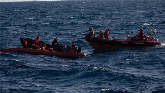 Mehrere Migranten sitzen in einem Boot im Mittelmeer während Rettungskräfte versuchen ihnen zu helfen.