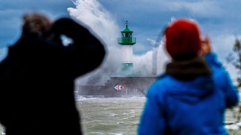 Menschen fotografieren Wellen der Ostsee, die bei einem schweren Sturmtief an den Leuchtturm auf der Ostmole peitschen.