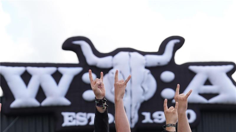 Metal-Fans zeigen vor dem Wacken Open Air-Logo die „Pommesgabel“, den Metal-Gruß, auf dem teilweise matschigen Festivalgelände des Wacken Open Airs (WOA). 