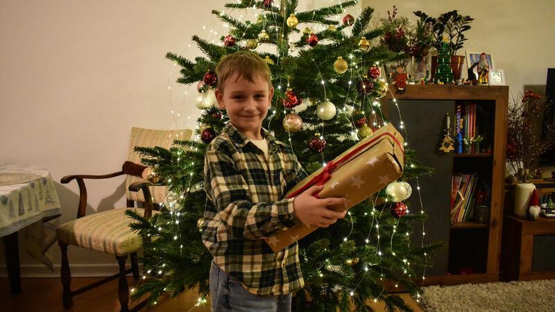 Misha Kushta steht vor einem Weihnachtsbaum und hält ein Geschnk in den Händen.