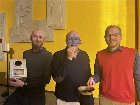 Mit diakonischem Blick: Pastor Volker Dieterich-Domröse guckt symbolisch durch das Kronenkreuz, das Diakonie-Logo. Links und rechts von ihm zeigen Töns Dittmer und Pastor Jan Wutkewicz zwei wichtige Arbeitswerkzeuge: Spendenbox und Klingelb