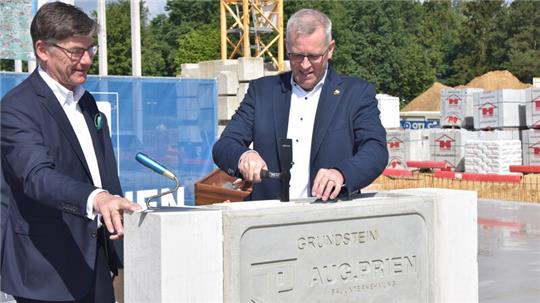 Mit einem Hammer versenkt Samtgemeinde-Bürgermeister Knut Willenbockel einen Ziegel in den Grundstein. Links von ihm steht Michael Groß, Geschäftsführer des Bauunternehmens Aug.Prien.