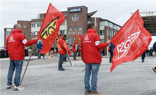 Mitarbeiter im Warnstreik stehen mit ihren Fahnen vor dem Haupteingang von Airbus in Hamburg-Finkenwerder. Die Gewerkschaft IG-Metall fordert einen Sozialtarifvertrag, in dem die Bedingungen für die von der Umstrukturierung betroffenen Beschäftigten geregelt werden. Arbeitsniederlegungen sind an allen betroffenen deutschen Standorten von Airbus und der Tochter Premium Aerotec Group geplant. Archivfoto: Markus Scholz/dpa