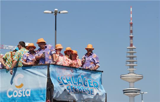 Musiktrucks mit zahlreichen Feiernden darauf fahren während des Schlagermoves auf dem Heiligengeistfeld. Im Hintergrund ist der Fernsehturm zu sehen. Foto: Georg Wendt/dpa