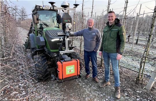 Obstbau setzt auf KI: Dr. Matthias Görgens im Gespräch mit Moritz Hentzschel vor einem Schlepper mit der Samson-Sensor-Box (von links) auf einer Versuchsplantage des Obstbauzentrums Esteburg in Moorende.