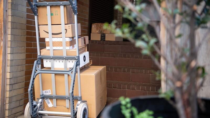 Es werden wieder mehr Pakete verschickt Pakete stehen auf einer Karre, während ein Paketzusteller Pakete ausliefert.