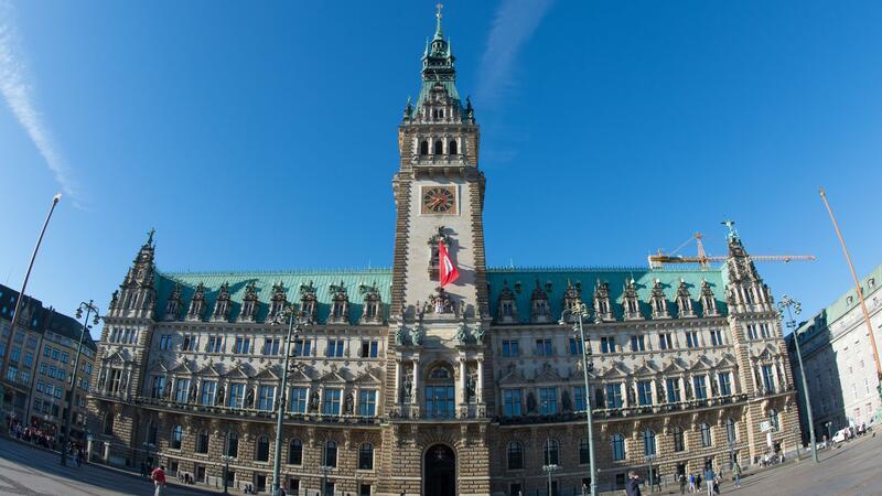 Passanten gehen in Hamburg bei strahlendem Sonnenschein über den Rathausmarkt.