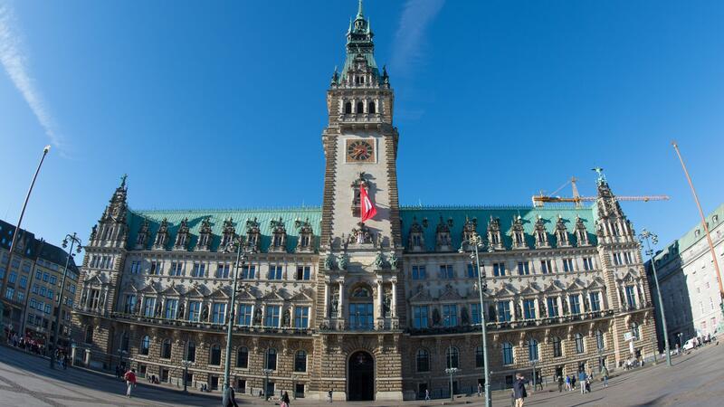 Passanten gehen in Hamburg bei strahlendem Sonnenschein über den Rathausmarkt.