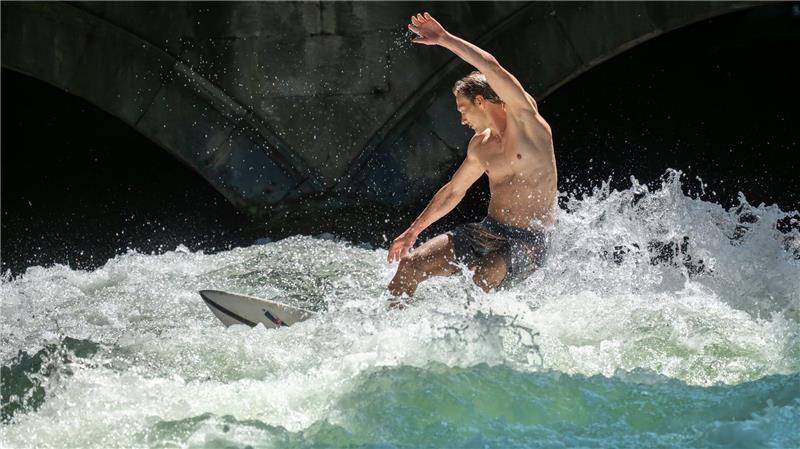 Patrick surft bei sommerlichen Temperaturen mit seinem Board auf der Eisbachwelle im Englischen Garten in München.