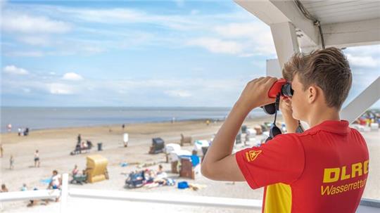 Peter von der DLRG-Wasserrettung steht auf einem Turm und beaufsichtigt mit einem Fernglas den Badestrand von Dangast.