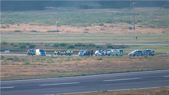 Polizeifahrzeuge und ein Follow-Me Wagen des Flughafen Köln-Bonn stehen auf einer Rollbahn. Aktivisten der Klima-Initiative Letzte Generation haben sich durch den Zaun geschnitten und als Protestaktion auf einer Zufahrt zu einer Start- und Landebahn festgeklebt.