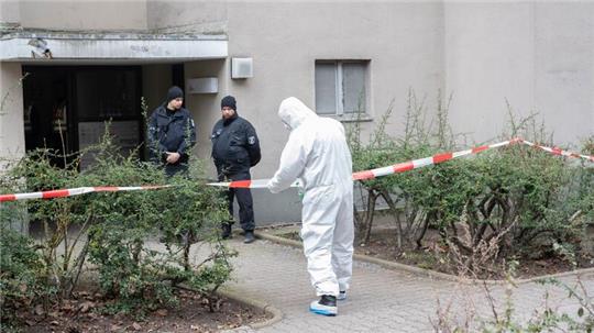 Polizisten stehen am Eingang eines Mehrfamilienhauses in Stadtteil Kreuzberg, während ein Polizist in Schutzanzug an einem Absperrband steht.