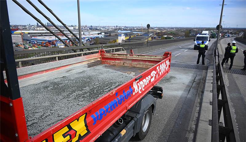 Polizisten stehen neben einem Laster, von dem Klimaaktivisten Kies auf die Fahrbahn gekippt hatten. Mehrere Klimaaktivisten haben den Verkehr auf der Hamburger Köhlbrandbrücke blockiert. Foto: Jonas Walzberg/dpa