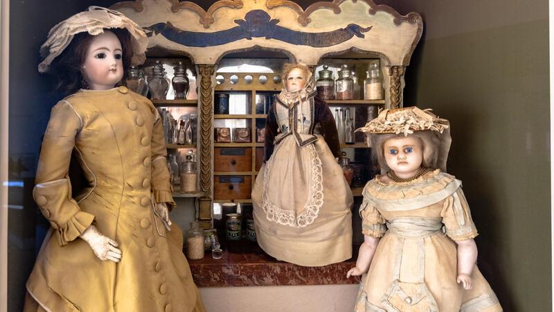 Von Porzellanpuppe bis Barbie: Auf Zeitreise im Puppenmuseum Falkenstein Drei große, in Röcken gekleidete Puppen stehen vor einem Miniaturregal mit Lebensmitteln.