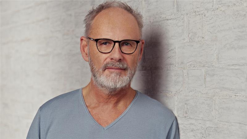 Reinhold Beckmann mit dunkler Brille und grauem Pullover vor einer hell getünchten Mauer 