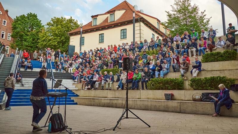 Barock in Stade: Vox Humana lädt zur musikalischen Zeitreise ein Chorleiterin Annegret Schönbeck steht am Ende der Treppen am Pferdemarkt und begleitet die auf den Stufen sitzenden Teilnehmer das gemeinsam Sing Along Open Air am Keyboard.