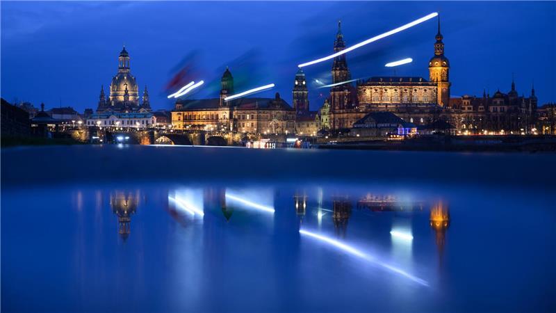 Scheinwerfer vorbeifahrender Radfahrer ziehen auf dem Elberadweg vor der Altstadtkulisse in Dresden Lichtspuren und spiegeln sich dabei in einer Pfütze.