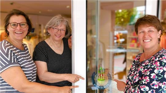 Sonja Gerken (von links), Carola Quast und Gabriela Strasdas zeigen auf das Symbol „Kinderinsel“, das am Harsefelder Mode- und Spielwarenhaus Quast klebt.