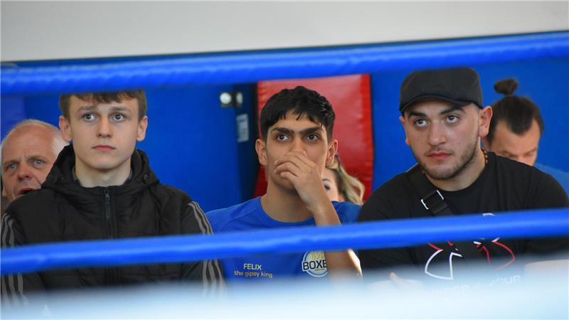 Drei junge Männer schauen einen Boxkampf von außerhalb des Boxrings.