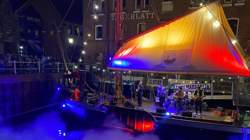 Fleth-Fest: Wie kommt das neue Buxtehuder Event an? Die Ewer-Bühne mit der Band Faela in rotes und blaues Scheinwerferlicht getaucht und Publikum entlang des Fleth-Geländers.