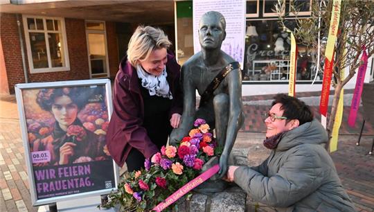 Symbole der Gleichberechtigung: Floristin mit Silke Liebau und Gleichstellungsbeauftragte Daniela Subei schmücken die Statue auf dem Platz an der T-Kreuzung "Im großen Sande/Lange Straße" zum Frauentag mit Nelken(von links).