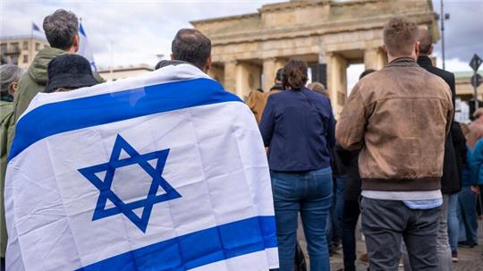 Teilnehmer der Kundgebung „Aufstehen gegen Terror, Hass und Antisemitismus – in Solidarität und Mitgefühl mit Israel“ stehen umhüllt in eine israelische Flagge vor dem Brandenburger Tor in Berlin.