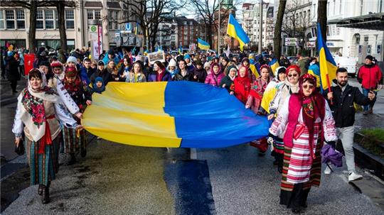 Teilnehmer einer Demonstration gegen den Krieg in der Ukraine ziehen am 24.02.2023 durch die Hamburger Innenstadt und tragen eine große Flagge.