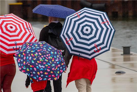 Touristen laufen mit Regenschirmen im Hafen. Die Meteorologen erwarten auch in den kommenden Tagen wechselhaftes Wetter. Foto: Stefan Sauer/dpa