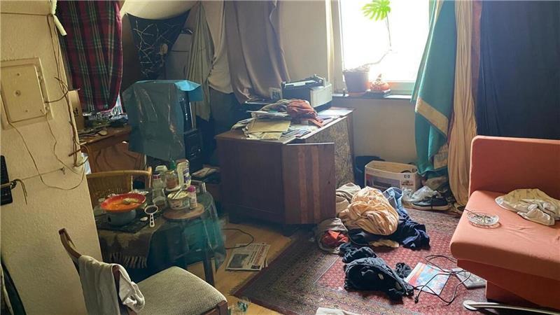Überall im Raum liegt schmutzige Kleidung, das Chaos ist allgegenwärtig; die Fenster sind mit Decken verhangen.