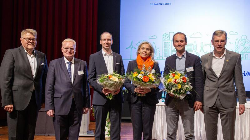 Volksbank Stade-Cuxhaven: Ein Auto-Freak erklärt Bankern das Verbrenner-Aus Ulrich Sievert (1. von Links) und Henning Porth (1. von rechts) vom Vorstand, Axel Lohse (2.v.l.) vom Aufsichtsrat, gratulieren auf der Bühne den Neuzugängen mit Blumensträußen: Daniel Hollung rückt in den Vorstand auf, Kim-Karen Liebig-Kusserow und Carsten Nickel sind neu im Aufsichtsrat.