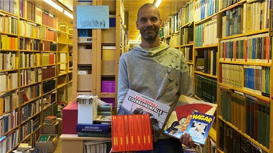Tobias Rothenberg steht mit einer Buchauswahl in den Händen zwischen den gefüllten Regalen von Hans-Uwe Hansens ehemaliger Privatbibliothek 
