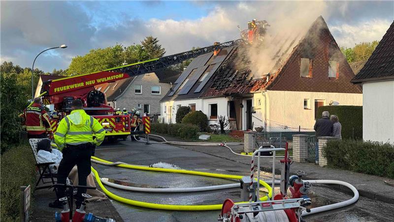 Hausbrand in Neu Wulmstorf: Bewohner erst gerettet, dann festgenommen Während die Feuerwehr das Haus löscht, beobachtet ein Mann im weißen Overall die Szene. Ein Polizist bewacht ihn.