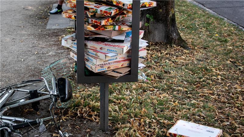 Zahlreiche Pizzakartons sind in einem Stahlgestänge für einen Mülleimer in einer Parkanlage gestapelt und liegen teilweise auf dem Rasen. 
