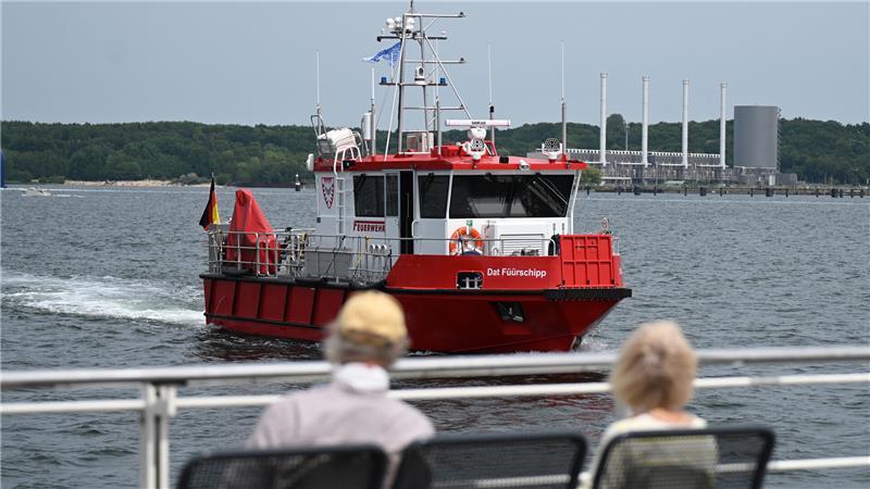Kieler Woche eröffnet: Sonne satt, wenig Wind Zwei Besucher schauen an der Kiellinie auf das Wasser, im Hintergrund fährt das Schiff „Dat Füürschipp“.