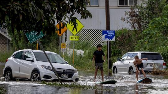 Zwei Jugendliche surfen in Waialua neben einem liegengebliebenen Fahrzeug im Hochwasser.