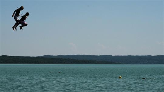 Zwei Jungen in Neoprenanzügen springen vom Sprungturm im Strandbad am Ammersee.