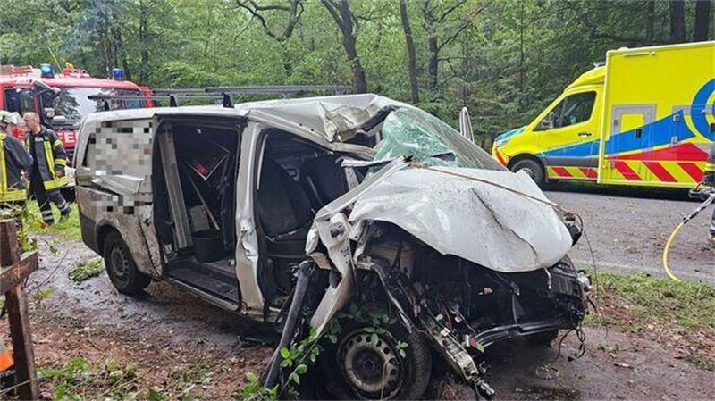 Ein zerstörtes Fahrzeug steht auf einem Gehweg, Rettungswagen und Feuerwehr sind im Hintergrund zu sehen.