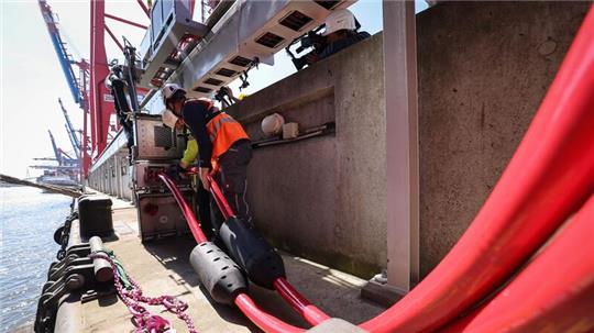 Zwei Techniker verbinden die Landstromkabel eines Containerschiffs mit einer verfahrbahren Steckdose entlang dem Kai.